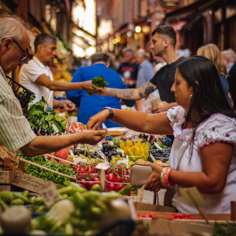 People browsing stalls at an outdoor food market, surrounded by fresh produce and local vendors