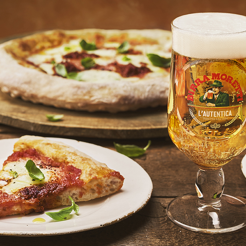 A slice of pizza on a plate next to a glass of Birra Moretti